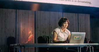 A woman sat at a table with a laptop. There is a screen with projected text behind her
