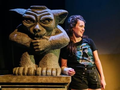 The performer is leaning against a gargoyle sculpture, wearing a cast on her right arm.