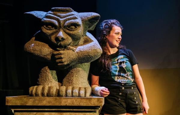 The performer is leaning against a gargoyle sculpture, wearing a cast on her right arm.