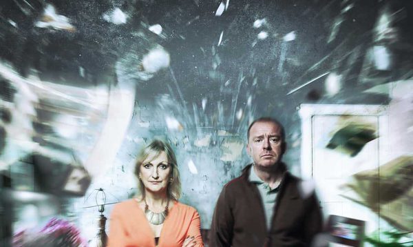 A man and woman stand facing forward. Around them, items from a living room explode around them.