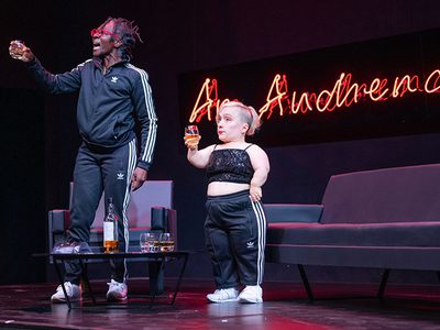 Two performers holding glasses up to toast, with a neon sign reading 'an audience' behind them