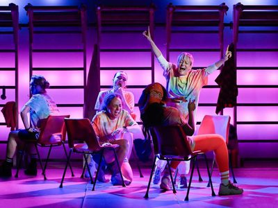 Group of performers wearing casual, brightly coloured clothing sit on stage against a backdrop of gym-style wall bars, illuminated by vivid pink and purple lighting. One performer stands behind with arms raised