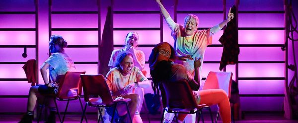 Group of performers wearing casual, brightly coloured clothing sit on stage against a backdrop of gym-style wall bars, illuminated by vivid pink and purple lighting. One performer stands behind with arms raised