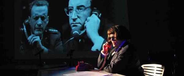 A woman sits at a table on the phone, behind her is a projected image of two men on phones