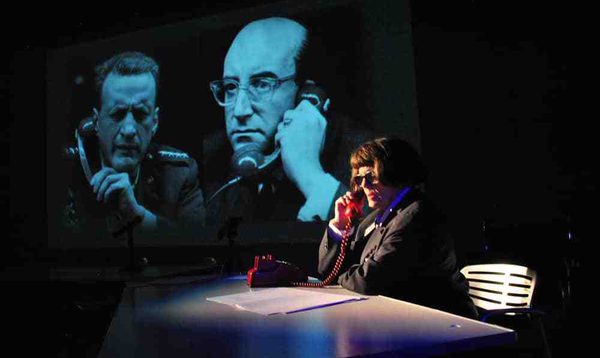 A woman sits at a table on the phone, behind her is a projected image of two men on phones