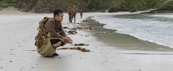 On a sand beach, a young soldier is crouched down, looking out to sea. In the distance, three more soliders stand by the edge of the sea.