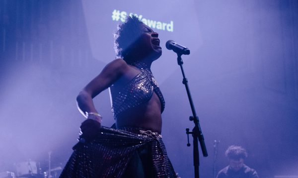 Beldina stands on-stage and sings into a microphone. The lighting is purple.