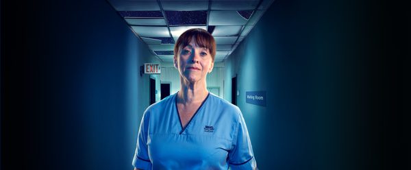 A nurse stands in a hospital corridor she is seems friendly yet strong. Her uniform is blue as is the lighting of the corridor behind her.