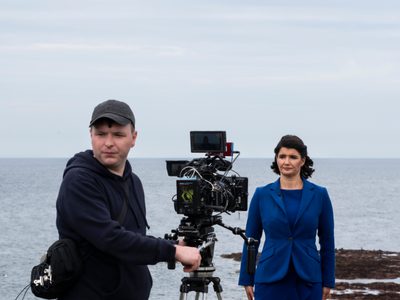 A woman in a blue suit stands in front of a camera, the camera operator looks over his shoulder, they are standing on a costal path