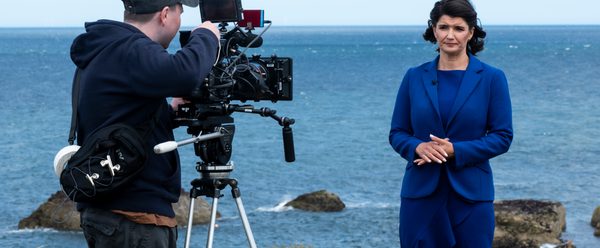 A newsreader in a blue suit stands in front of a camera, the camera operator looks down the lens, they are standing on a cliffside costal path with the ocean in the background