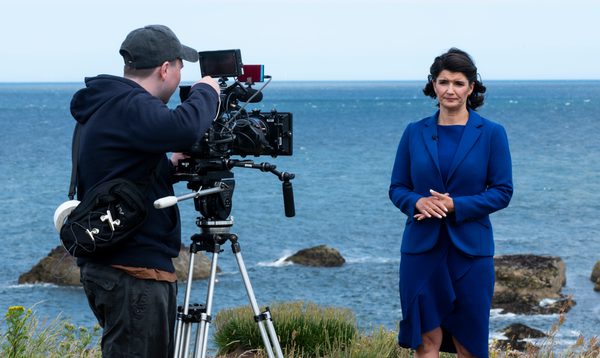 A newsreader in a blue suit stands in front of a camera, the camera operator looks down the lens, they are standing on a cliffside costal path with the ocean in the background