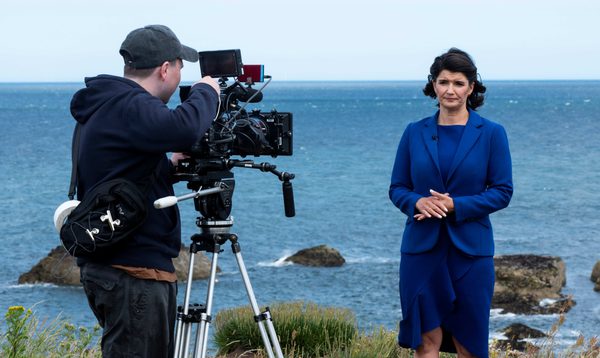 A newsreader in a blue suit stands in front of a camera, the camera operator looks down the lens, they are standing on a cliffside costal path with the ocean in the background