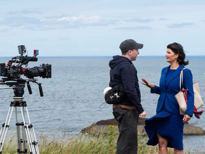A woman in a blue suit and a man in a black hoodie standing talking to each other on a costal path in front of a video camera
