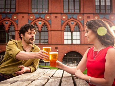 A man and woman sit at a picnic bench, they clink their beer glasses