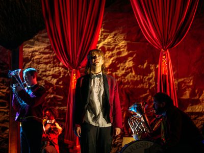 Keli (Liberty Black) stands looking upwards. Two musicians are behind her playing brass instruments. Red curtains are draped behind them. The lighting is warm and golden.