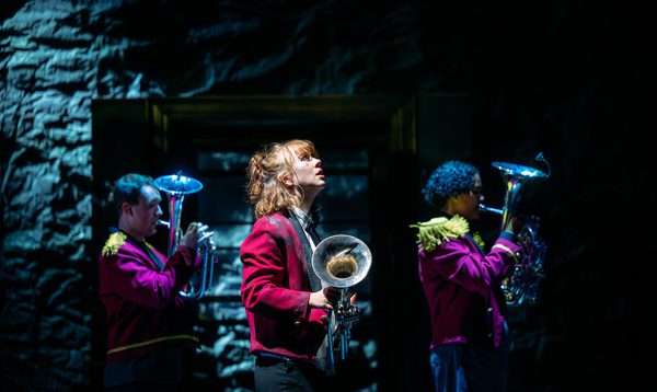 Liberty Black stands looking upwards holding her trombone. Two other performers playing trombones stand behind her.
