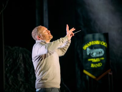 Phil Mckee stands conducting with a baton. His eyes are closed.
