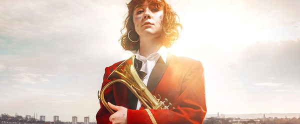 KELI lead actor, Liberty Black stands against scottish landscape holding a brass horn. she has paint smeared on her face