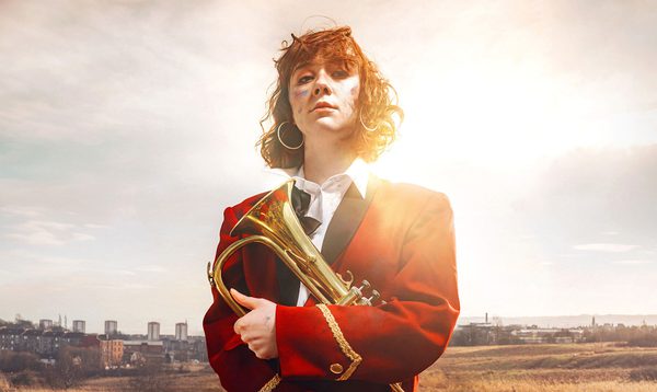 KELI lead actor, Liberty Black stands against scottish landscape holding a brass horn. she has paint smeared on her face