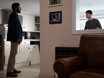 Sani Mamood and Atta Yaqub stand facing each other having an intense conversation in a kitchen living room.