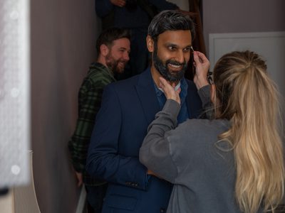 A blonde makeup artist touches up Atta Yaqub's make up. Atta is smiling. A member of the crew is walking past in the background.