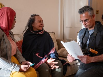 Kal Sabir looks kneels in front of Farah Ahmed and Kanchan Raval. They sit on a couch looking warmly at him while he looks intently at a script.