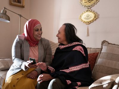 Kanchan Raval and Farah Ahmed sit in a living room on the couch smiling at each other. Farah is holding mobile phone.
