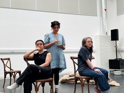 Two actresses sit on chairs. One actress stands. They are in a rehearsal room and all look concerned.