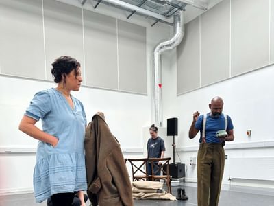 Actress Nicole Cooper stands firm. She is holding a brown jacket. Ramesh has a teacup, he seems distracted.