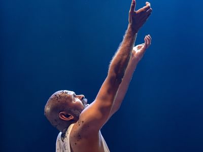 Ramesh Meyyappan as Lear. His hands are raised up as though talking to a higher power. He wears a simple white vest top. The background is a midnight blue colour.