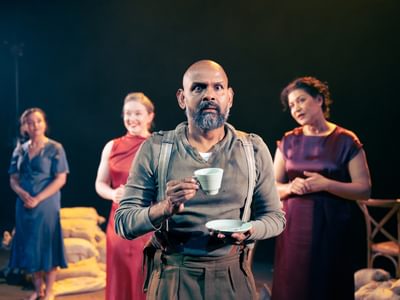 Lear is front and centre. He holds a tea cup and saucer, his expression is concerned and scared. In the background, actresses Amy Kennedy and Draya Maria smile and seem excited. Also in the background is Nicole Cooper who looks at Lear with a curious expression.
