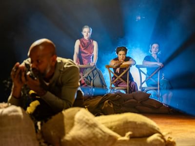 Lear inspects compost from a sack, he looks terrified. In the background yet in-focus are the three actresses who watch on. Draya's expression is soft but Nicole glares at Lear. A white-blue light shines behind the women giving them a dreamlike glow.