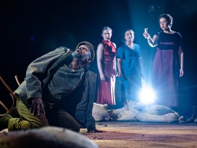 Lear kneels on the ground, he looks up to the sky in terror. The three actresses watch him from behind, accompanied by the white-blue glow. Nicole has some concern in her expression and hold a small object up.