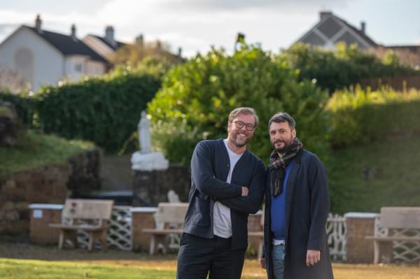 Damian Barr and James Ley are standing in green space at Carfin Grotto, with benches and a statue behind them.