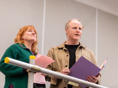 A woman and a man stand together on a high platform, looking out. They are both holding scripts.