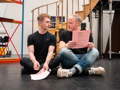 Two men sit on the ground with their legs crossed, looking at one another, holding their scripts.