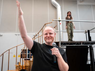 A man holds a microphone in his left hand with his right arm raised. A woman is standing on a platform behind him, singing into a microphone on a stand. 