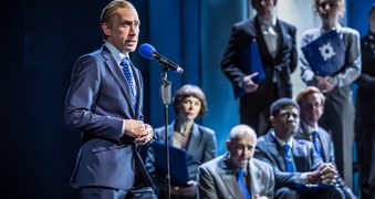 Sandy Grierson stands in front of a microphone, he has his hands clasped and wears a suit. Seven actors are behind him. They watch intently.