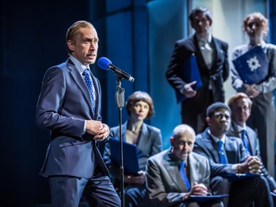 Sandy Grierson stands in front of a microphone, he has his hands clasped and wears a suit. Seven actors are behind him. They watch intently.