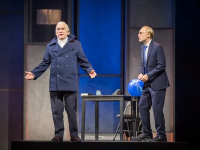 Actors Sandy Grierson and Brian Cox stand on stage. Sandy wears a suit and fidgets with his fingers, he is nervous and looks at Brian in admiration. Brian wears a powdered wig and a suit which is the same shade of navy as Sandy's but Brian's suit is more 18th century in style.