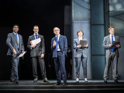 Sandy Grierson stands on stage with four other actors behind him. Sandy appears to be talking and the actors listen intently, they seem excited; driven; and important.