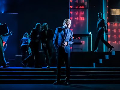 Sandy Grierson stands centre-stage, he looks to the side. Behind him five actors can be seen. Three are walking and two whisper to each other.