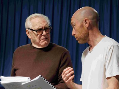 Sandy Grierson and Brian Cox talk to each other. Brian holds a script and looks befuddled. Sandy is intently explaining something.