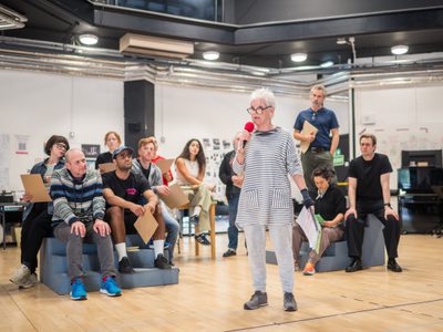 Ann Louise Ross stand in the centre of the room, holding a microphone. Ten actors sit on chairs behind her.