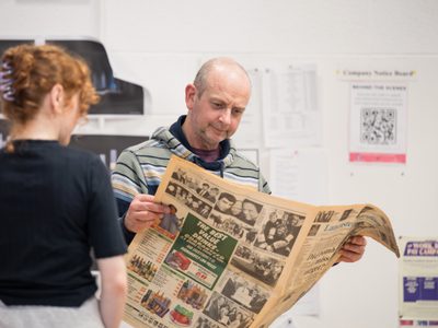 Actor David Mara reads a newspaper. A female actor watches him, she has her back to the camera.