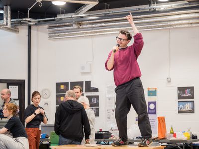 Actor Lawrence Smith stands on a table he is wearing a mulberry jumper and holds a microphone. He appears to be singing while five other actors look the other way.