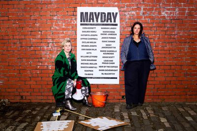 Two women are standing next to a red brick with a poster that reads MAYDAY with a list of participating artists. They have protest signs and a megaphone.