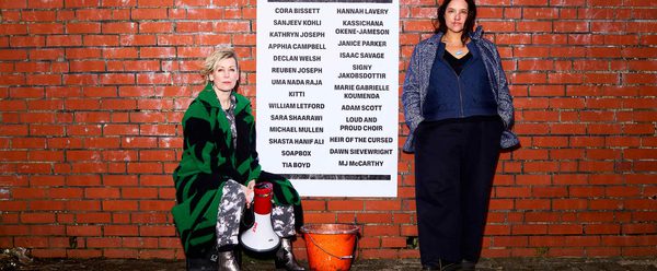 Two women are standing next to a red brick with a poster that reads MAYDAY with a list of participating artists. They have protest signs and a megaphone.