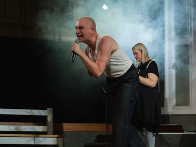 Tom Rowan leans forward singing into a microphone with an angry expression. A blonde woman stands behind him. There is haze an a soft white light behind them both.