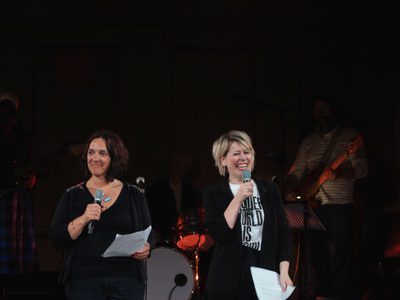 Hannah Lavery and Cora Bisset stand holding microphones and pieces of paper. They are both smiling. A band are behind them in the shadows.